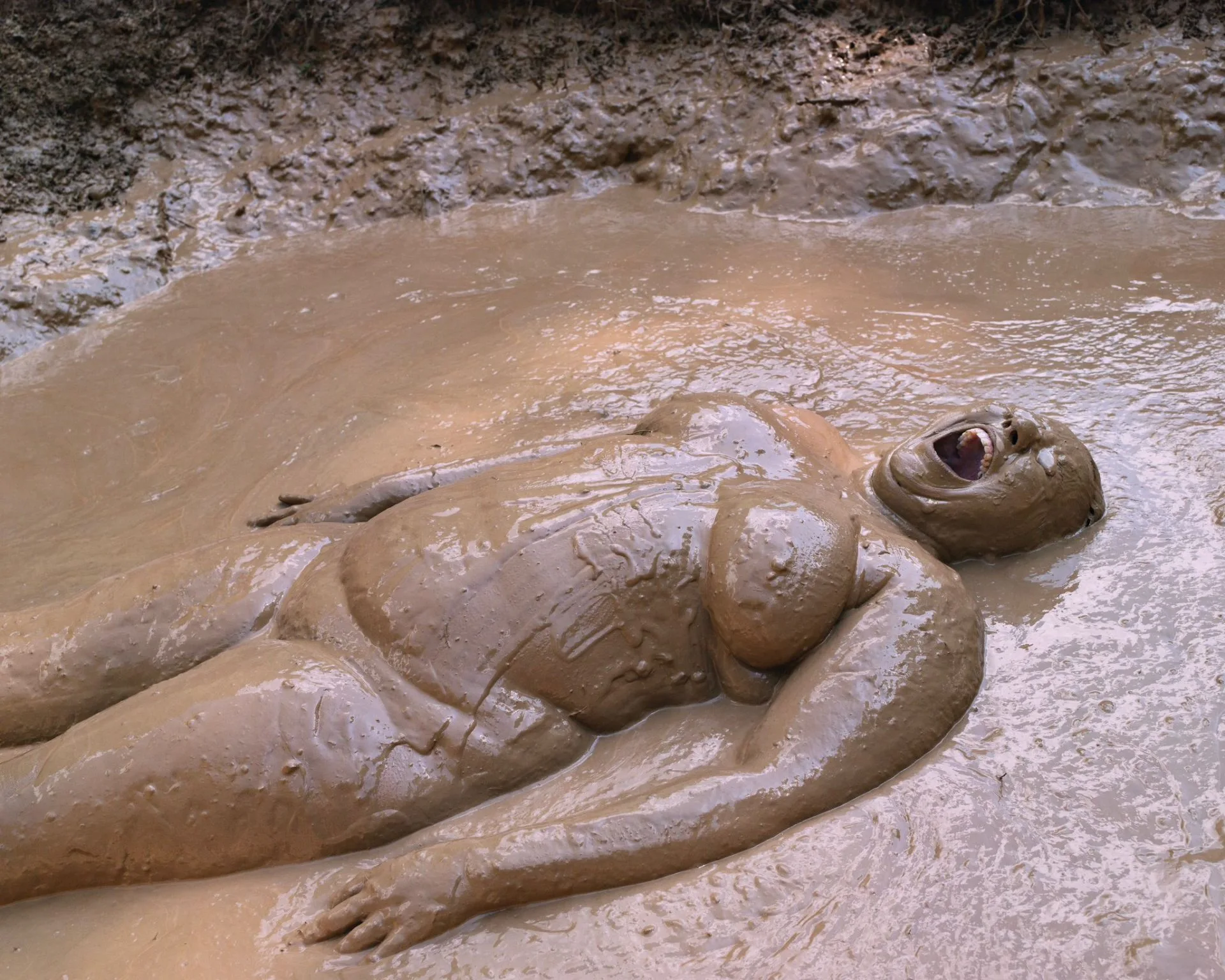 Une personne allongée sur le dos, entièrement couverte de boue, dans une fosse remplie d'eau boueuse, par Lucas Foglia.