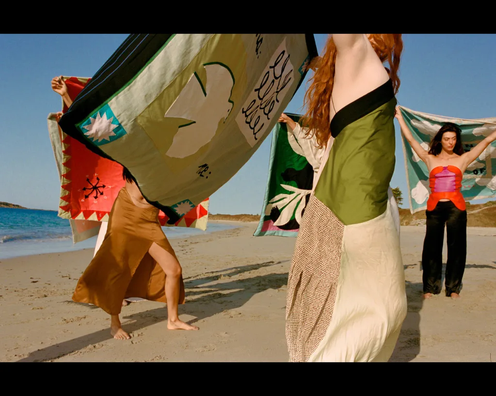 Sur une plage, des femmes en robes amples brandissent au vent de grands tissus cousus de motifs, sous un ciel bleu.
