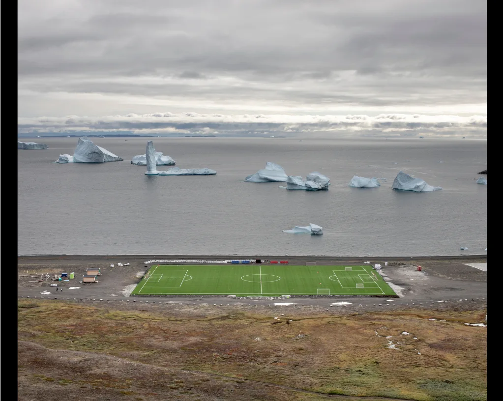 Terrain de football au gazon synthétique vert vif, au bord d'une mer grise parsemée d'icebergs, par Marco Zorzanello.