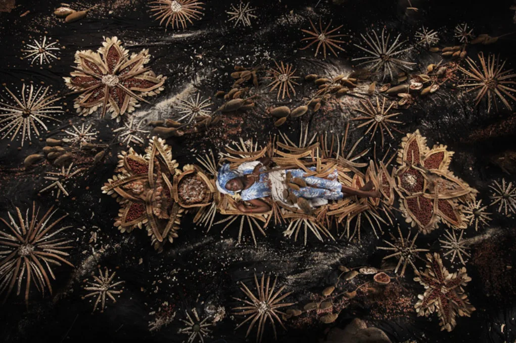 Vue d'en haut, une personne en tenue bleue allongée au centre de rosaces de cosses et de fibres sur fond noir, par Nicolas Henry.