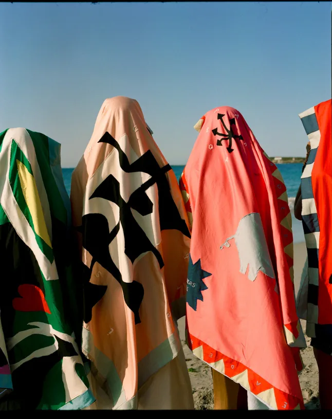 Quatre personnes de dos sur une plage, la tête couverte de grands tissus colorés ornés de motifs découpés, face à la mer.