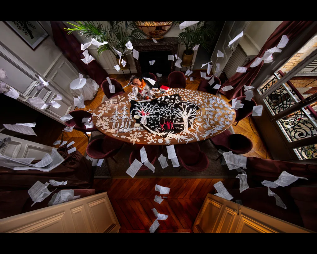 Vue plongeante sur une salle de réunion : une grande table couverte d'arbres en papier découpé, des feuilles volant dans les airs.