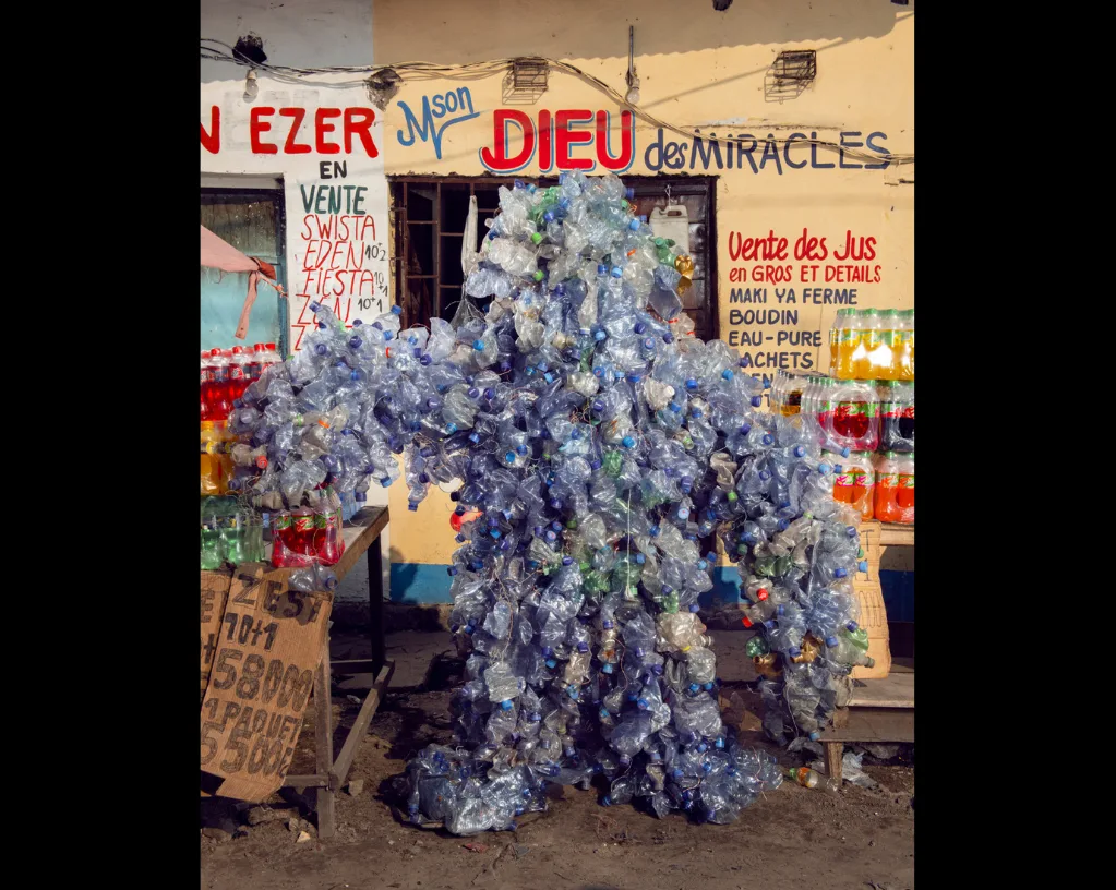 Personnage entièrement recouvert de bouteilles d'eau en plastique vides, devant une buvette aux enseignes peintes en rouge.