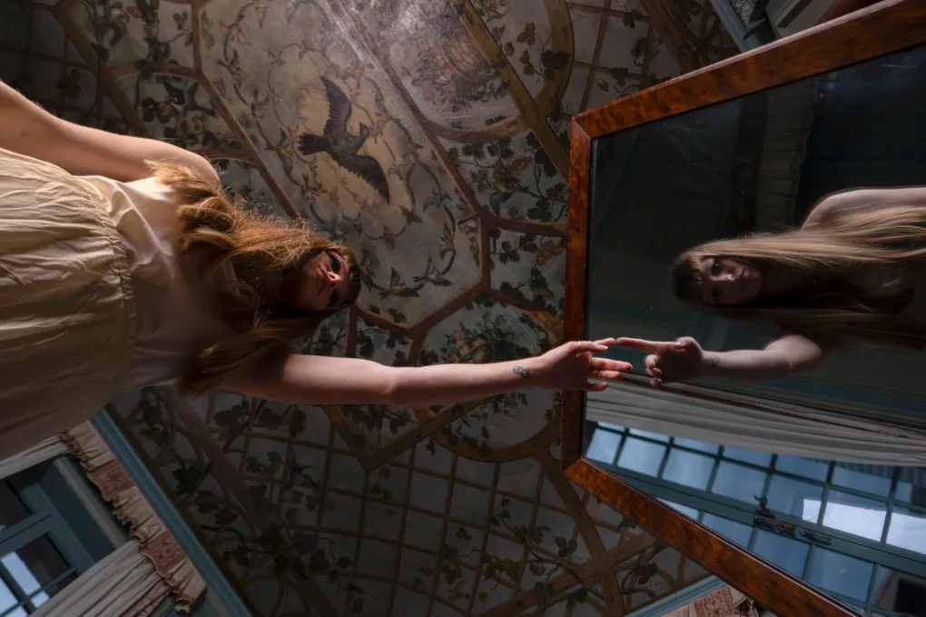 Une femme aux longs cheveux, en robe beige, tend la main vers son reflet dans un grand miroir, sous un plafond peint d'oiseaux.