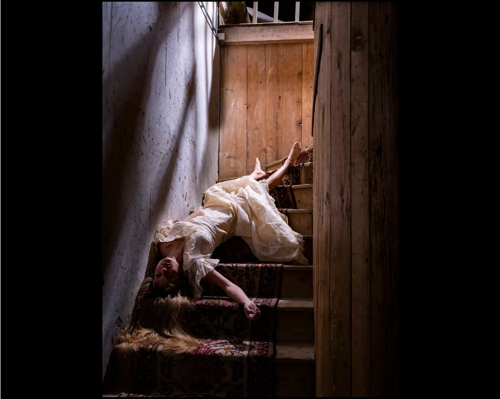 Une femme en robe blanche gît à la renverse sur un escalier recouvert d'un tapis, ses longs cheveux répandus sur les marches.