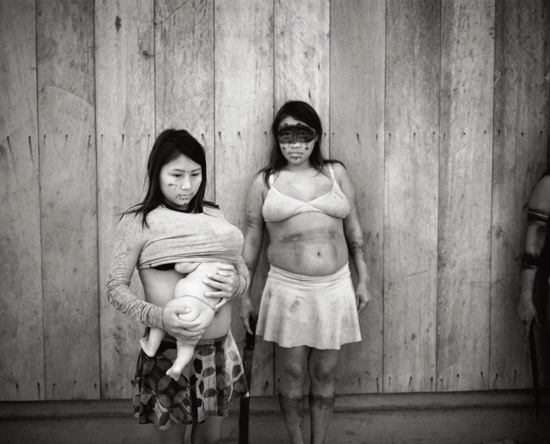 Deux femmes au visage peint, l'une tenant un bébé, debout devant une paroi en planches, en territoire yanomami, en noir et blanc.