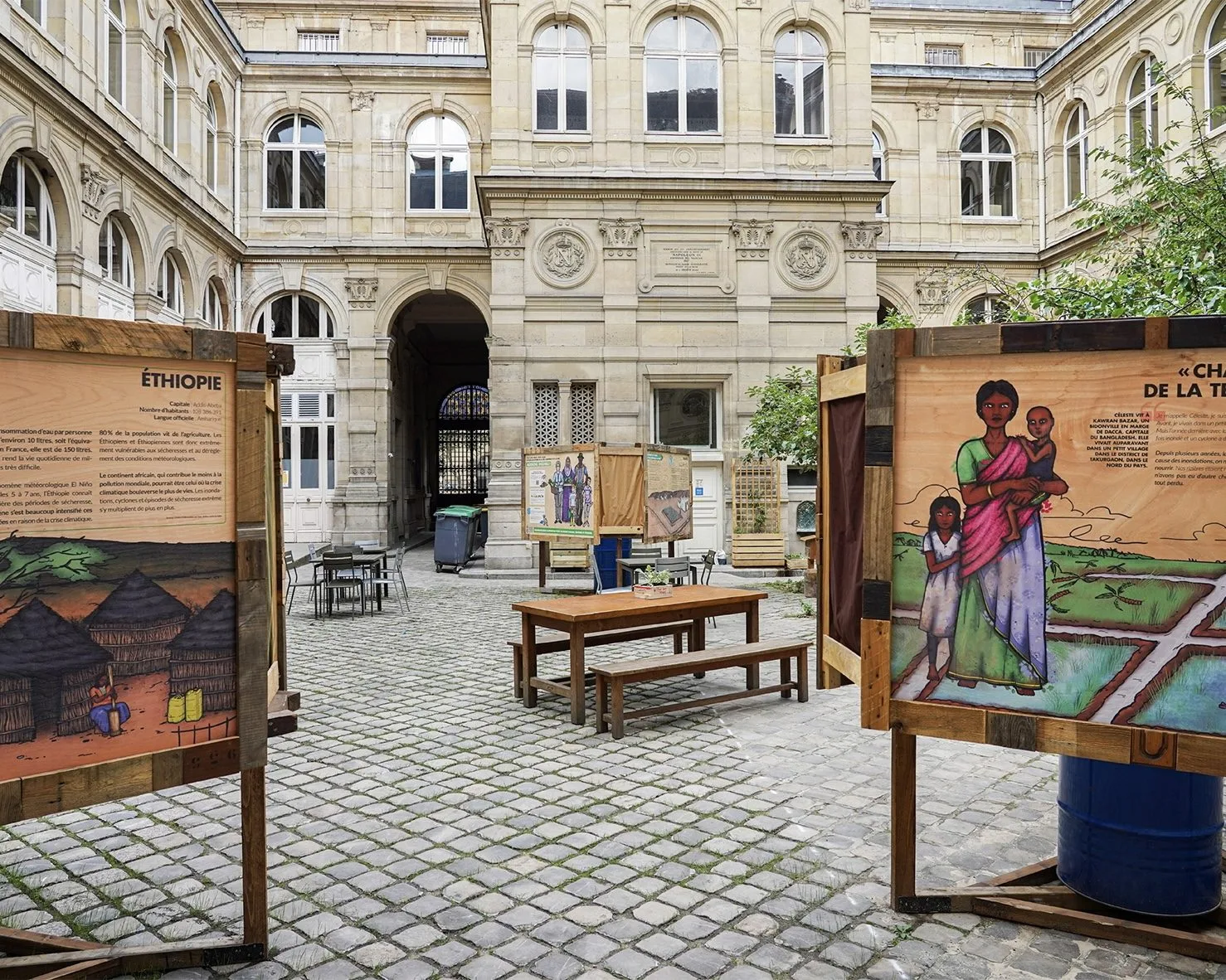 Panneaux illustrés en bois installés dans la cour pavée d'un bâtiment ancien, à l'Académie du Climat, autour d'une table en bois.