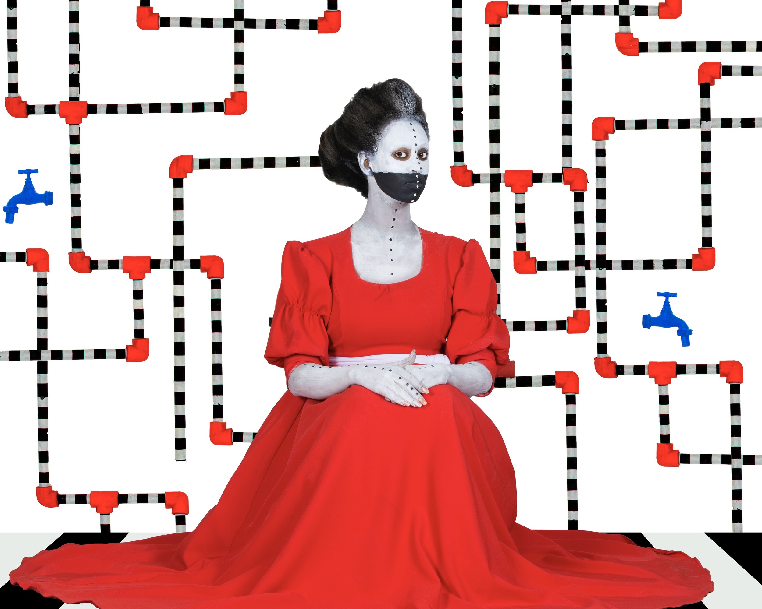 Une femme au visage peint en blanc et masquée de noir, en robe rouge, assise devant un réseau de tuyaux noir et blanc et des robinets bleus.