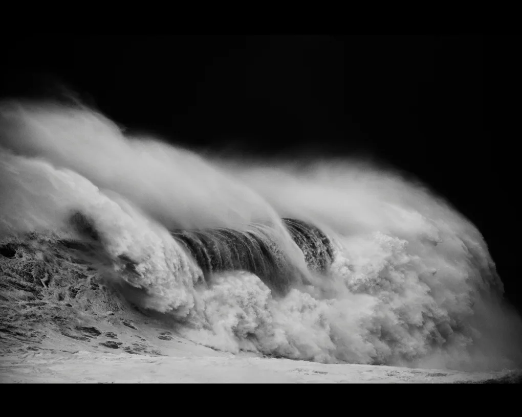 En noir et blanc, une vague déferle de gauche à droite dans un nuage d'embruns, sur un fond entièrement noir, par Alessandro Puccinelli.