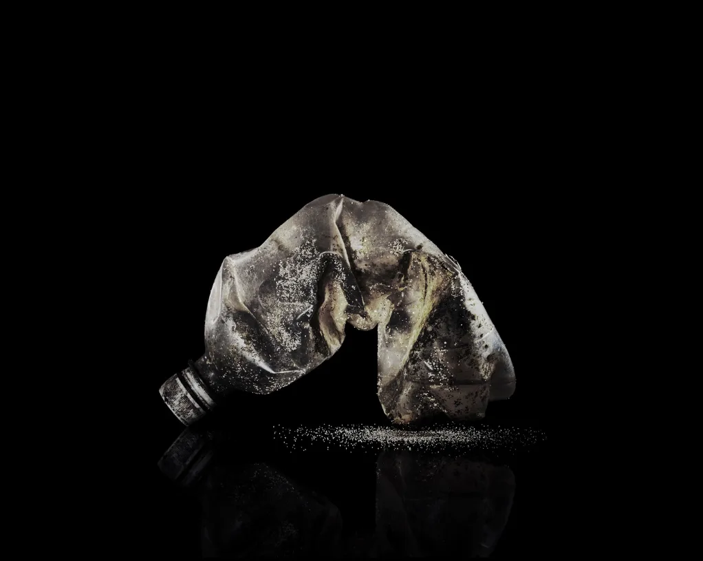 Une bouteille en plastique écrasée, usée et salie, posée sur un fond noir où brillent quelques grains de sable, par Alessandro Puccinelli.