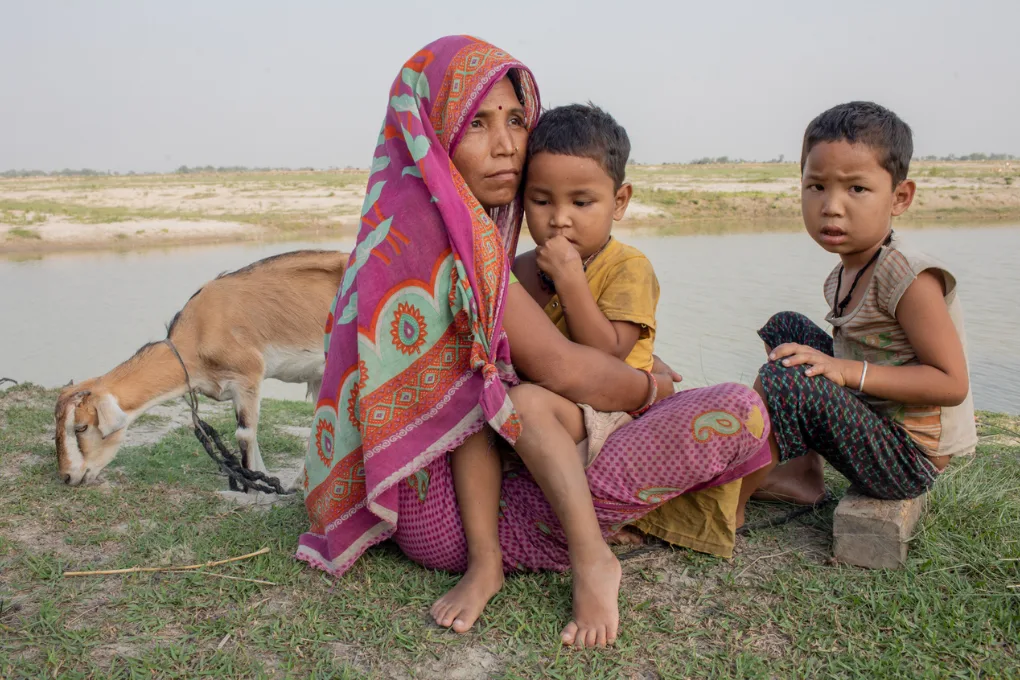 Une femme en voile rose serre un enfant contre elle, un autre garçon assis à côté, une chèvre broute au bord de la rivière Kosi.