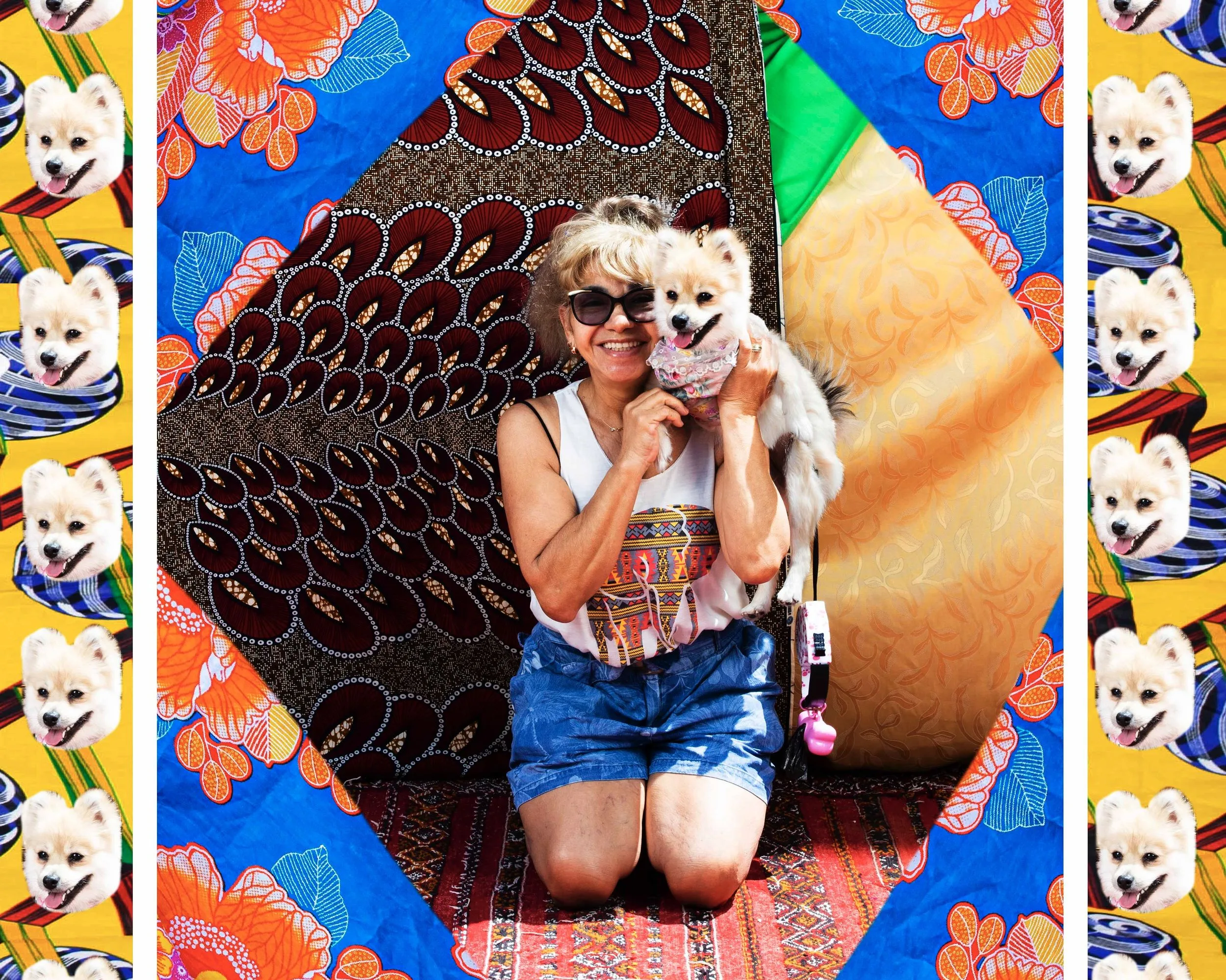 Une femme en lunettes de soleil, à genoux sur un tapis devant des tissus imprimés colorés, serre contre elle un petit chien blanc.