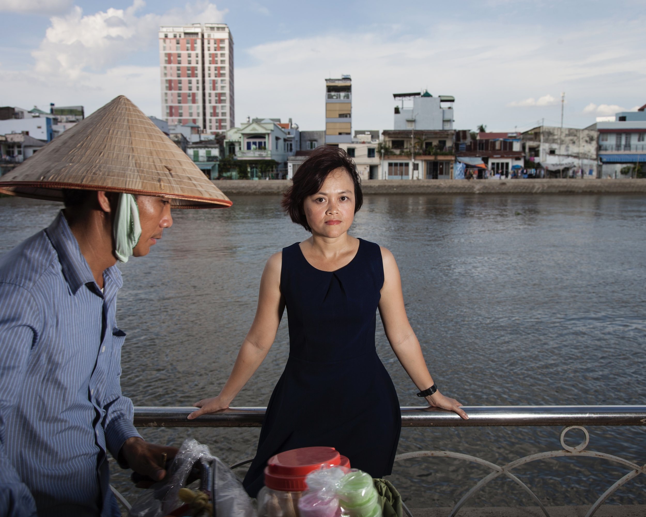 Une femme en robe bleu marine pose devant une rambarde au bord d'un fleuve en ville ; au premier plan, un vendeur au chapeau conique.