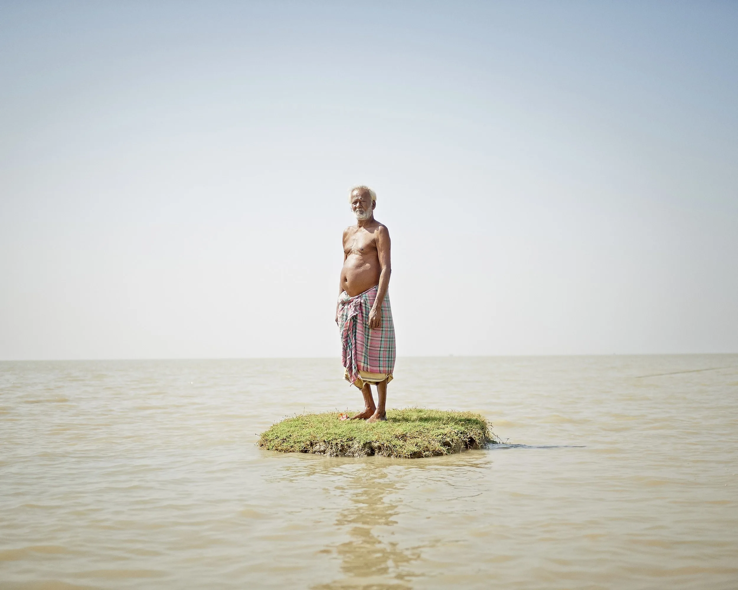 Un homme âgé, torse nu et vêtu d'un pagne à carreaux, debout sur un minuscule îlot d'herbe au milieu d'une eau boueuse, par Daesung Lee.