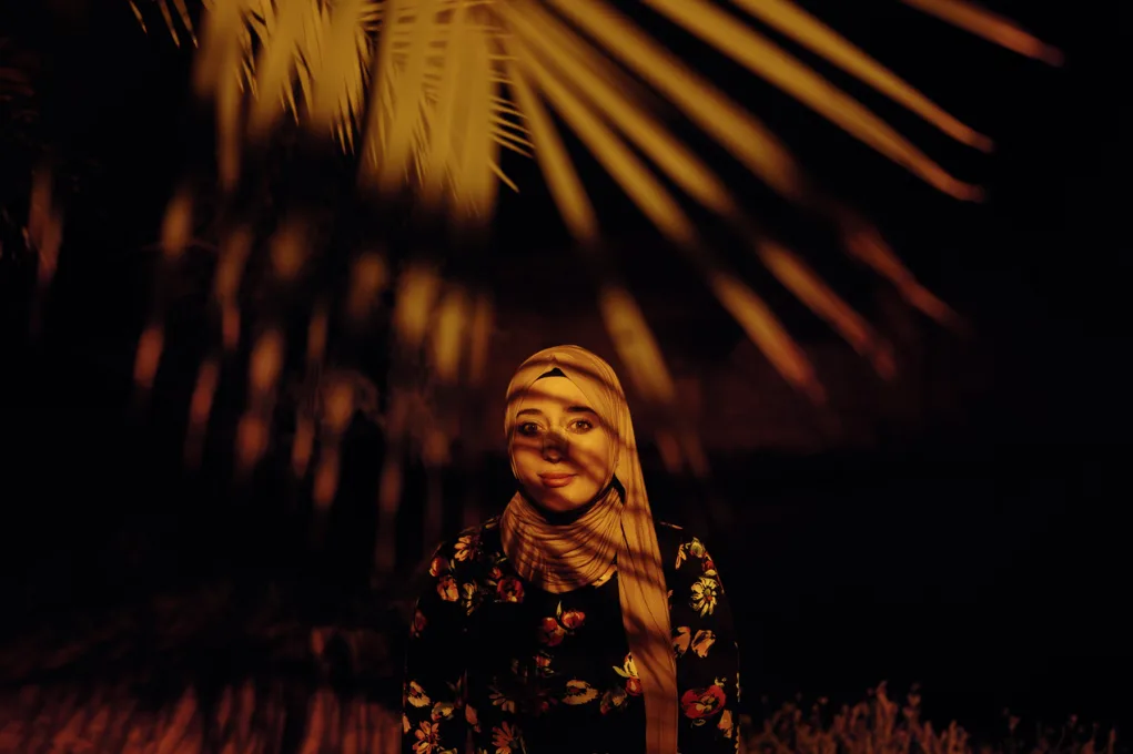 De nuit, une femme au foulard beige sourit sous des palmes éclairées d'une lumière orangée, leurs ombres striant son visage.