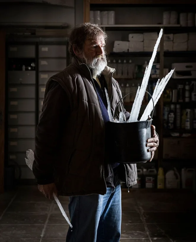 Un homme barbu en gilet tient un seau noir rempli de longues pointes blanches, comme des stalactites de glace, dans un local sombre.
