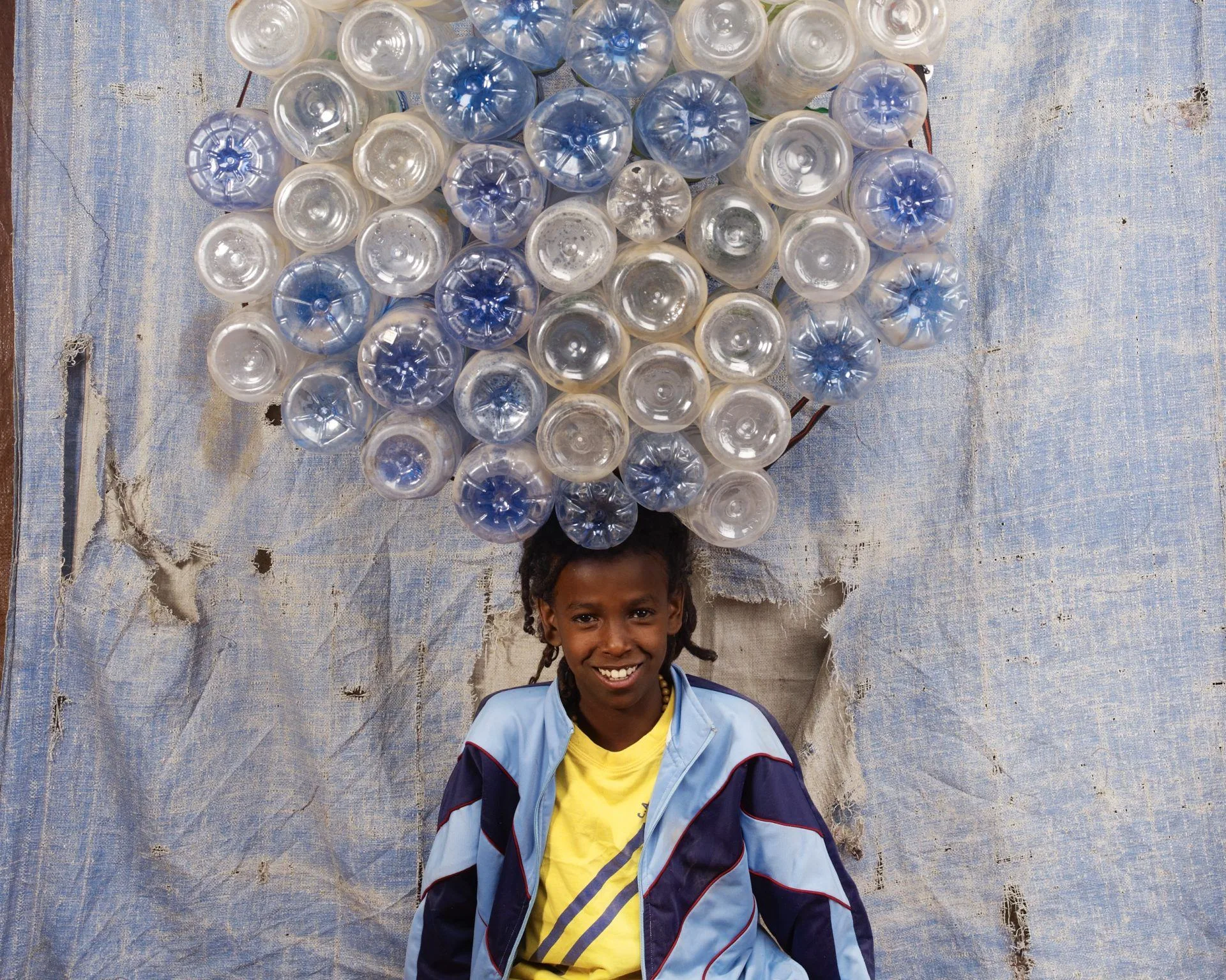 Un jeune rasta sourit, une couronne de bouteilles en plastique bleues et transparentes sur la tête, devant une bâche, par Floriane de Lassée.