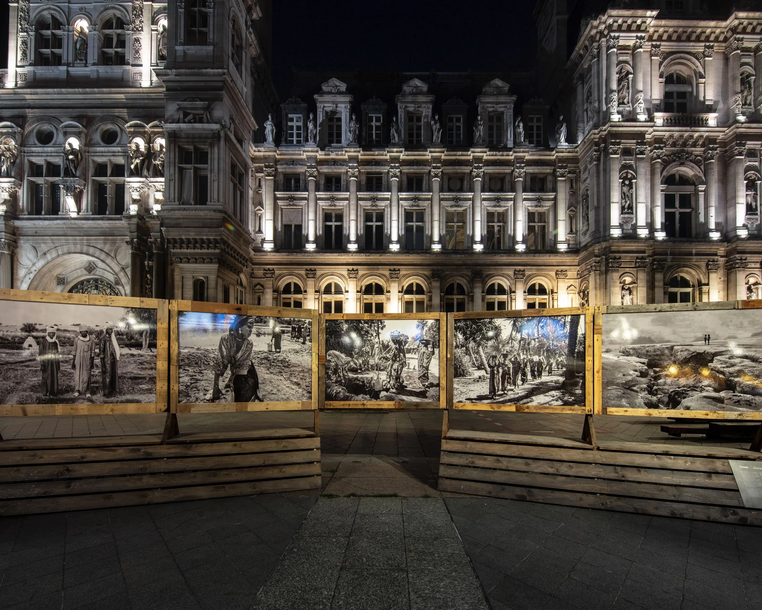 De nuit, une rangée de tirages noir et blanc sur panneaux de bois devant la façade illuminée de l'Hôtel de Ville de Paris.