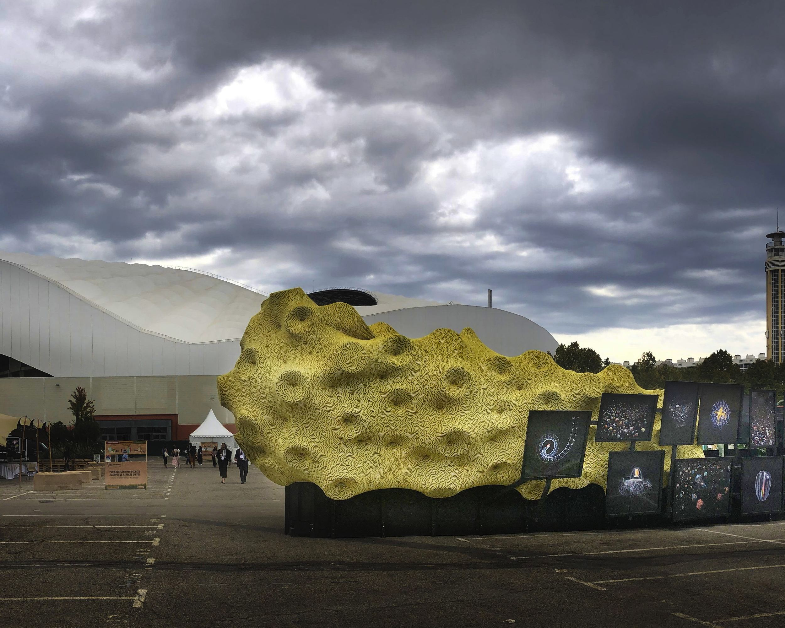 Sculpture jaune alvéolée de Jérémy Gobé, « Cerveau de Neptune », bordée de tirages de plancton, sous un ciel d'orage.