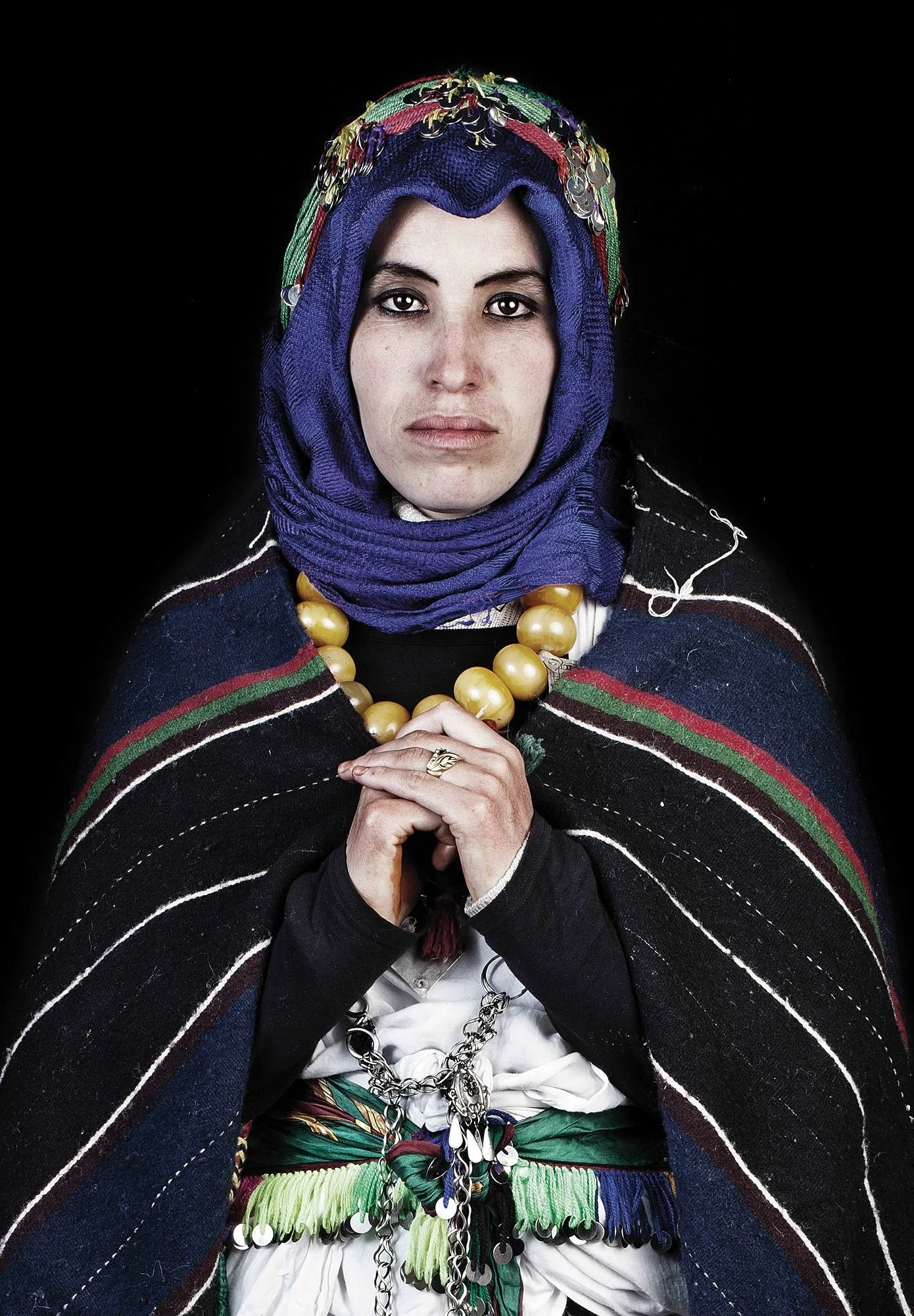 Portrait de Leila Alaoui : une femme au foulard bleu, collier de grosses perles dorées et cape rayée, mains jointes sur fond noir.