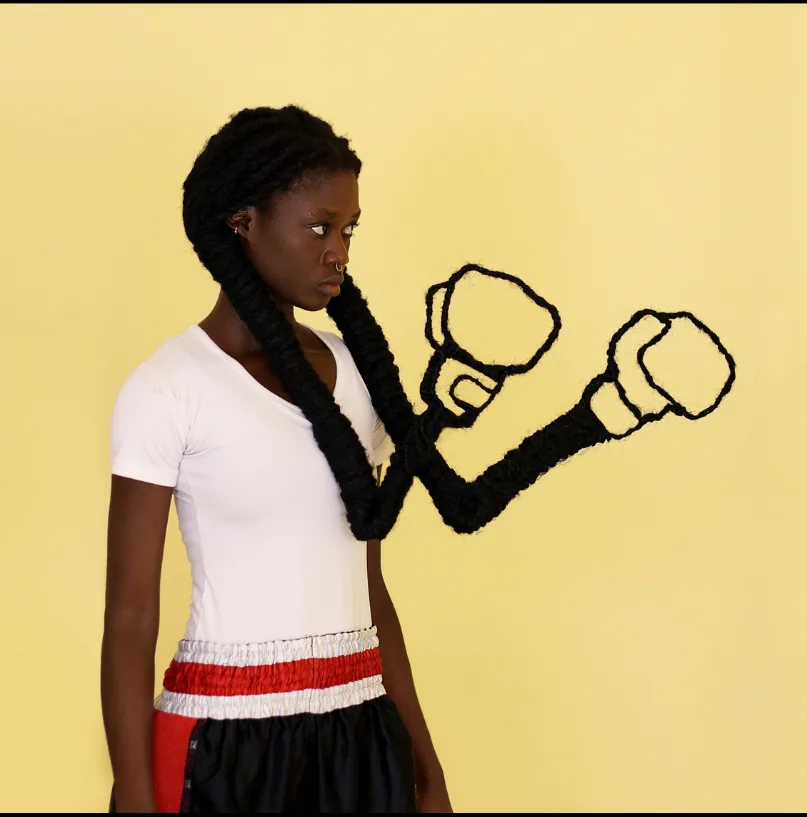 Une jeune femme sur fond jaune, dont les longues tresses sculptées forment deux bras terminés par des gants de boxe, œuvre de Laetitia Ky.