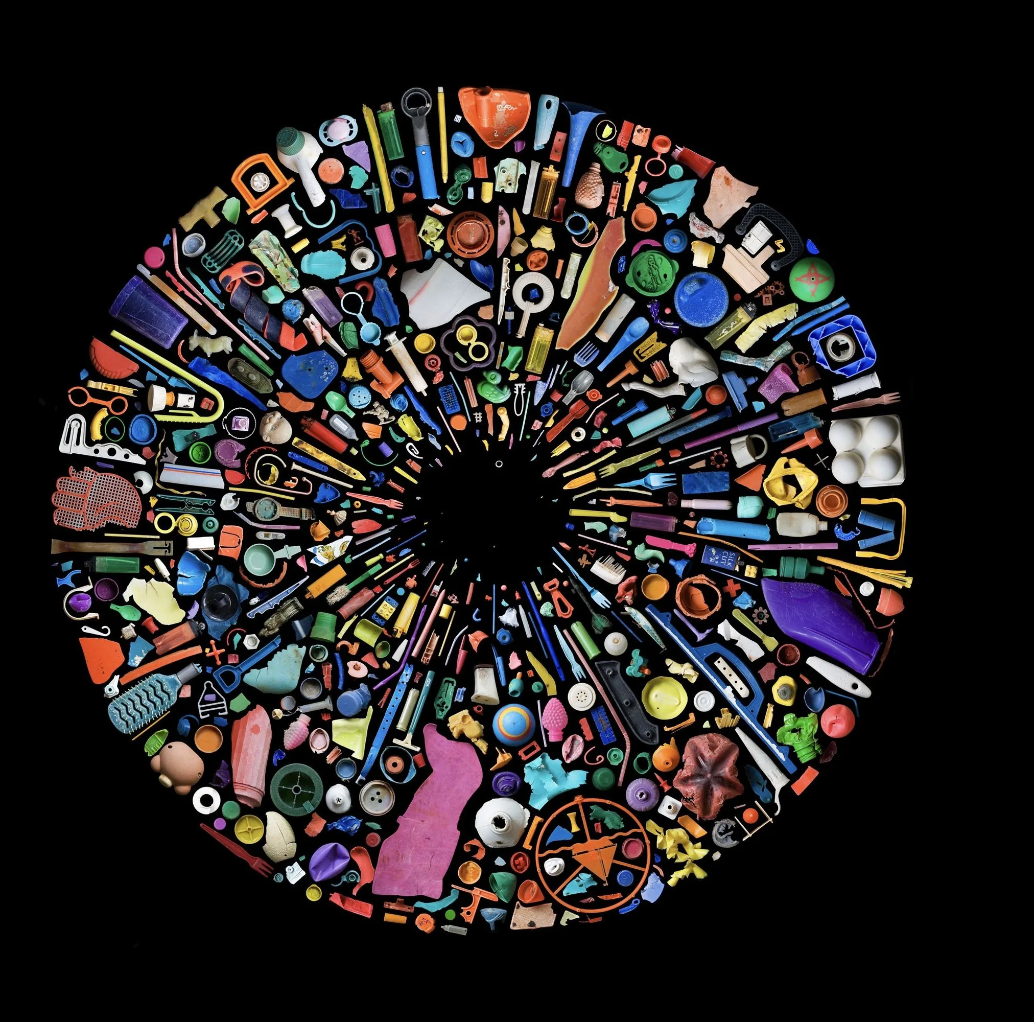 Cercle de centaines de débris de plastique colorés disposés en rayons sur fond noir, autour d'un centre vide, par Mandy Barker.