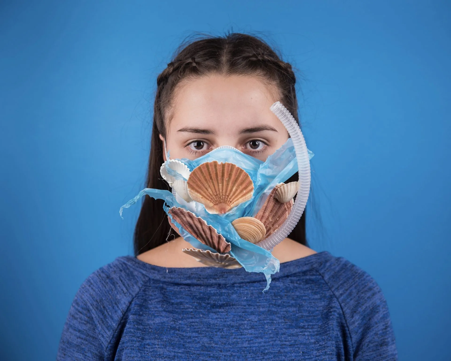 Portrait d'une jeune femme sur fond bleu, portant un masque chirurgical orné de coquilles Saint-Jacques et d'un tuyau annelé.