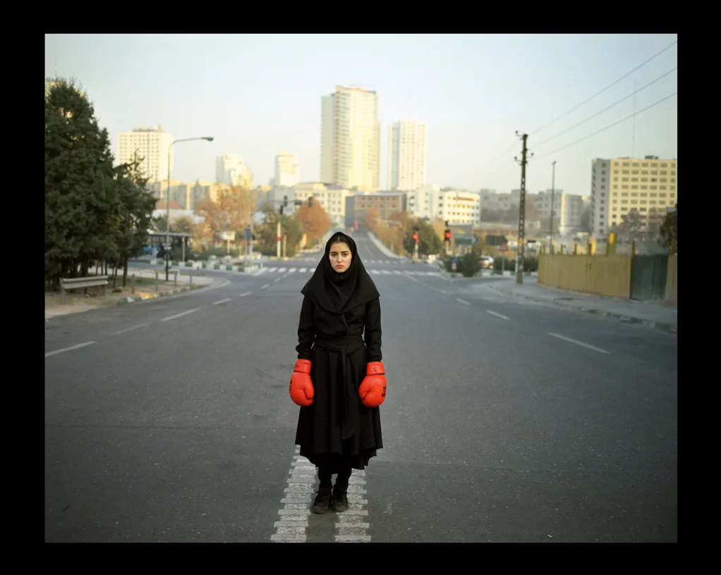 Une jeune femme en voile et manteau noirs, gants de boxe rouges aux mains, debout au milieu d'une avenue vide bordée de tours.