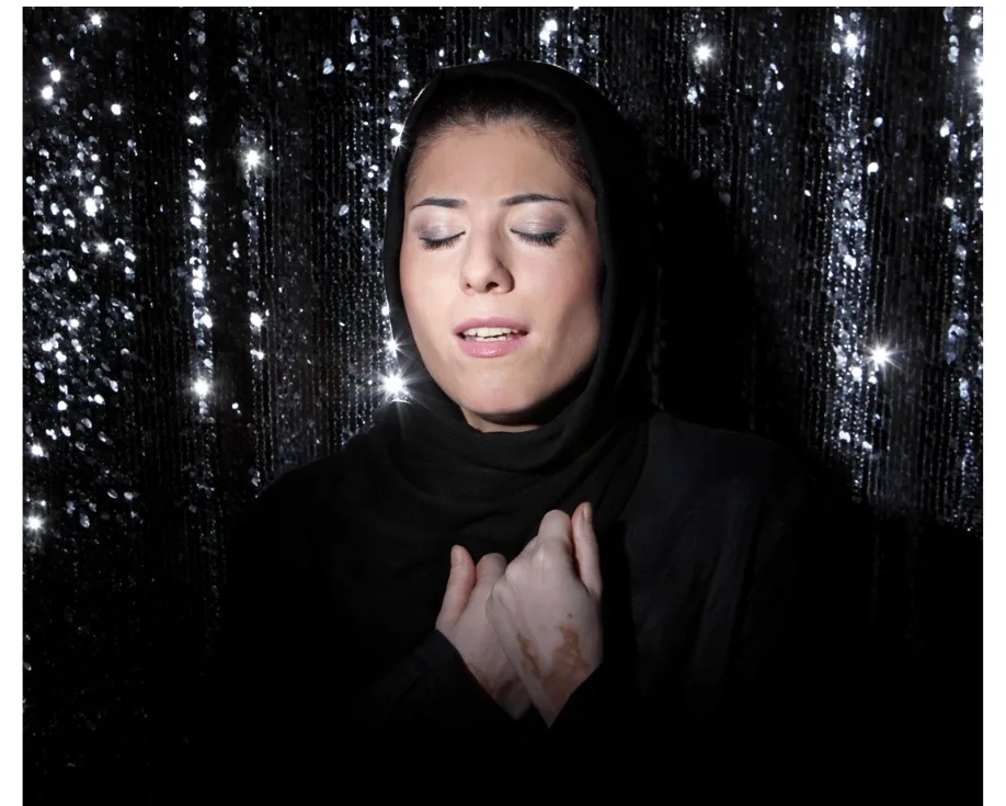 Une femme voilée de noir chante les yeux fermés, les mains jointes sur la poitrine, devant un rideau scintillant, par Newsha Tavakolian.