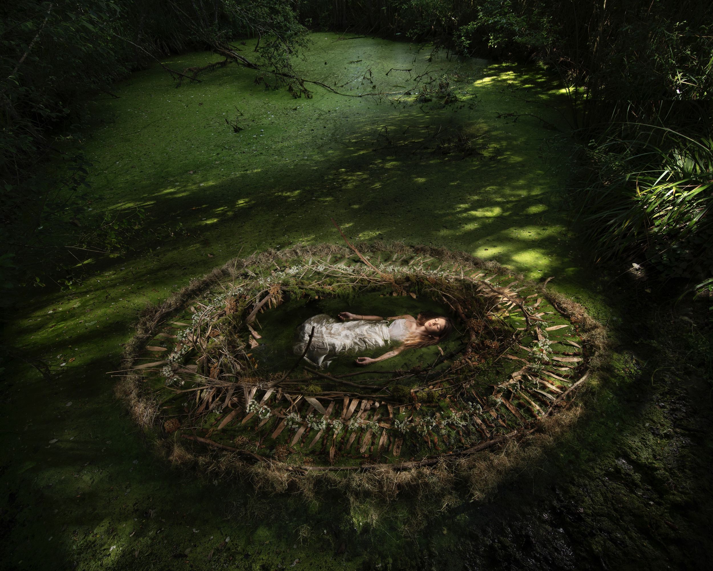 Une femme en robe claire allongée au centre d'un grand nid circulaire de branches, posé sur une mare verte en sous-bois, par Nicolas Henry.