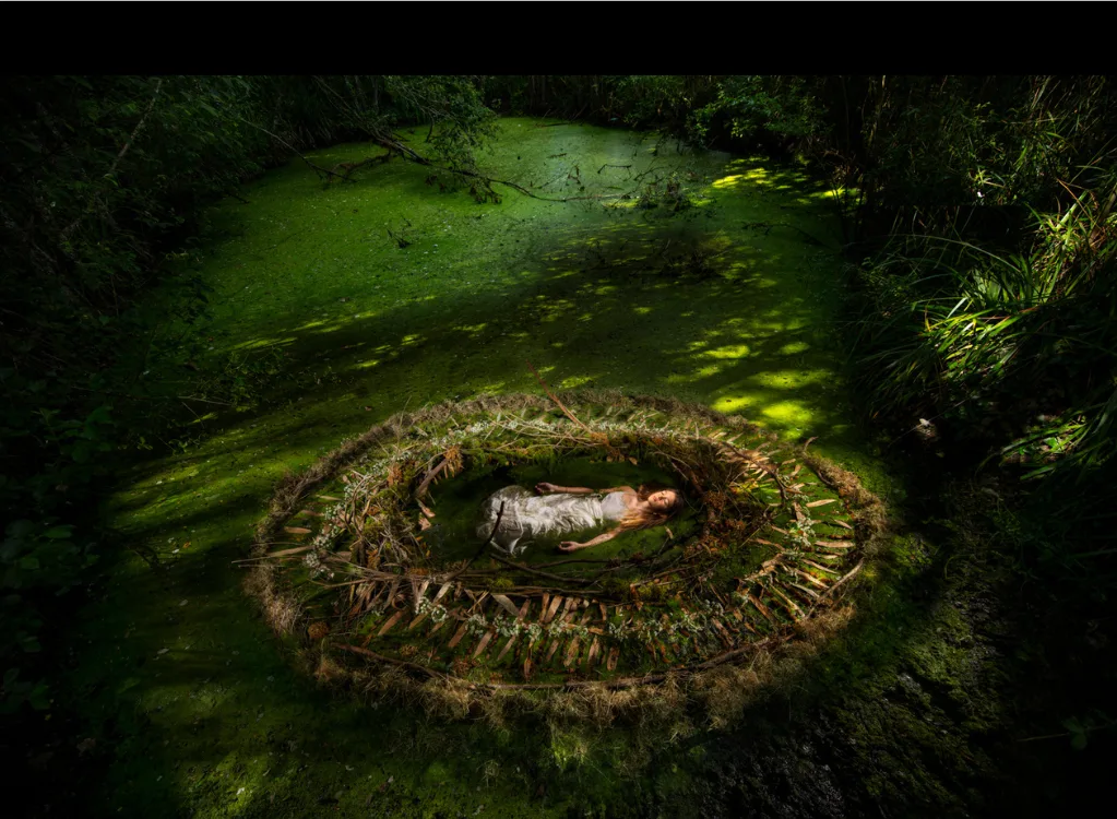 Une femme en robe claire allongée dans un nid circulaire de branchages, posé sur une mare couverte de lentilles d'eau, par Nicolas Henry.