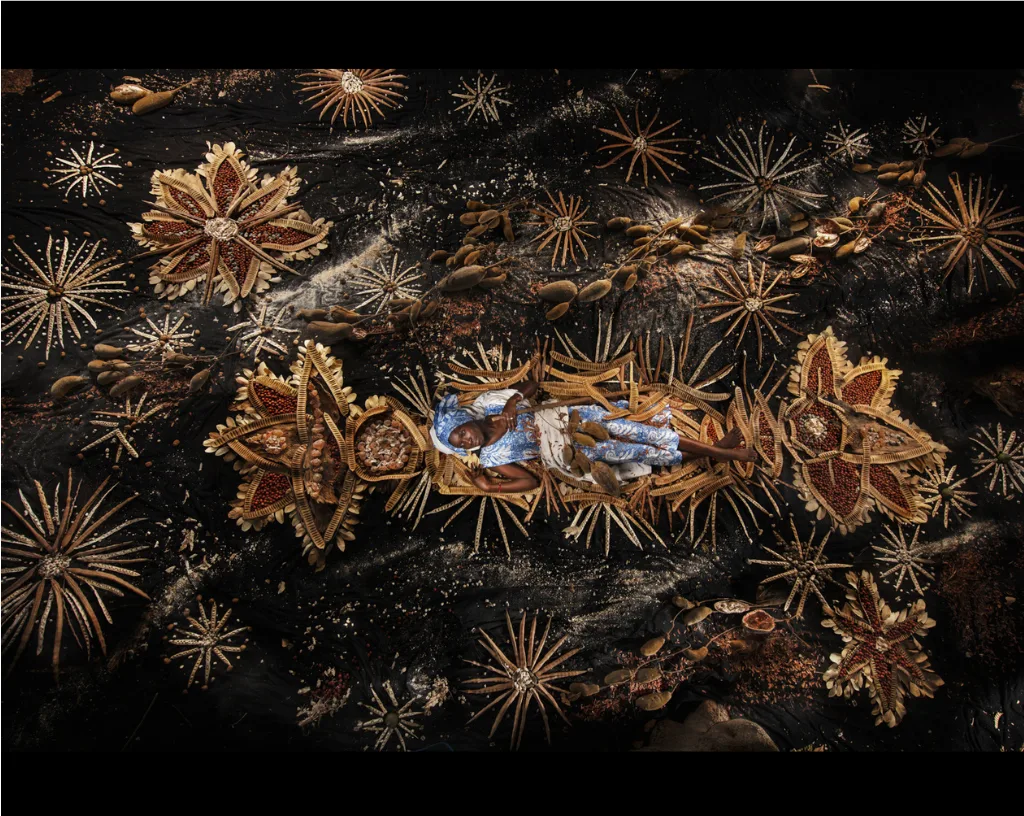 Une personne en tenue bleue allongée sur fond noir, au cœur d'un décor de cosses, de fibres et de graines en étoiles, par Nicolas Henry.