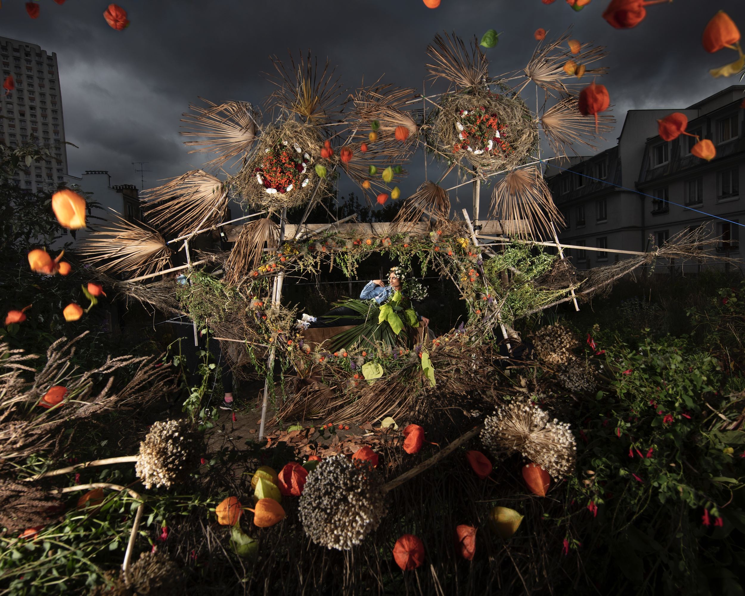Grande cabane de paille, d'herbes sèches et de lanternes orange sous un ciel d'orage en ville, une personne assise au centre, par Nicolas Henry.
