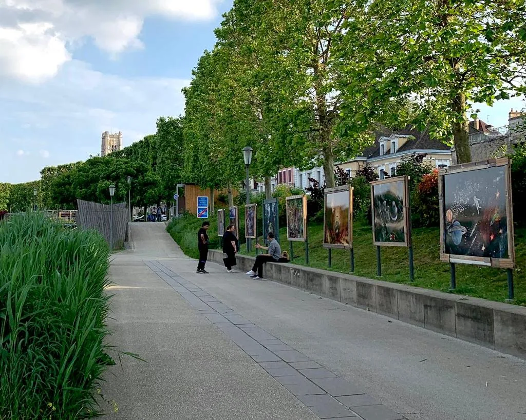 Grands tirages colorés de Nicolas Henry alignés sur des pieds métalliques le long d'une promenade arborée, des passants assis sur le muret.