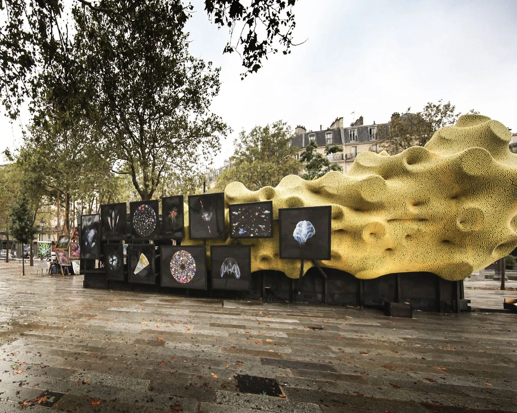 Tirages de Mandy Barker et de plancton sur fond noir, alignés devant la grande sculpture jaune de Jérémy Gobé, sur un trottoir mouillé.