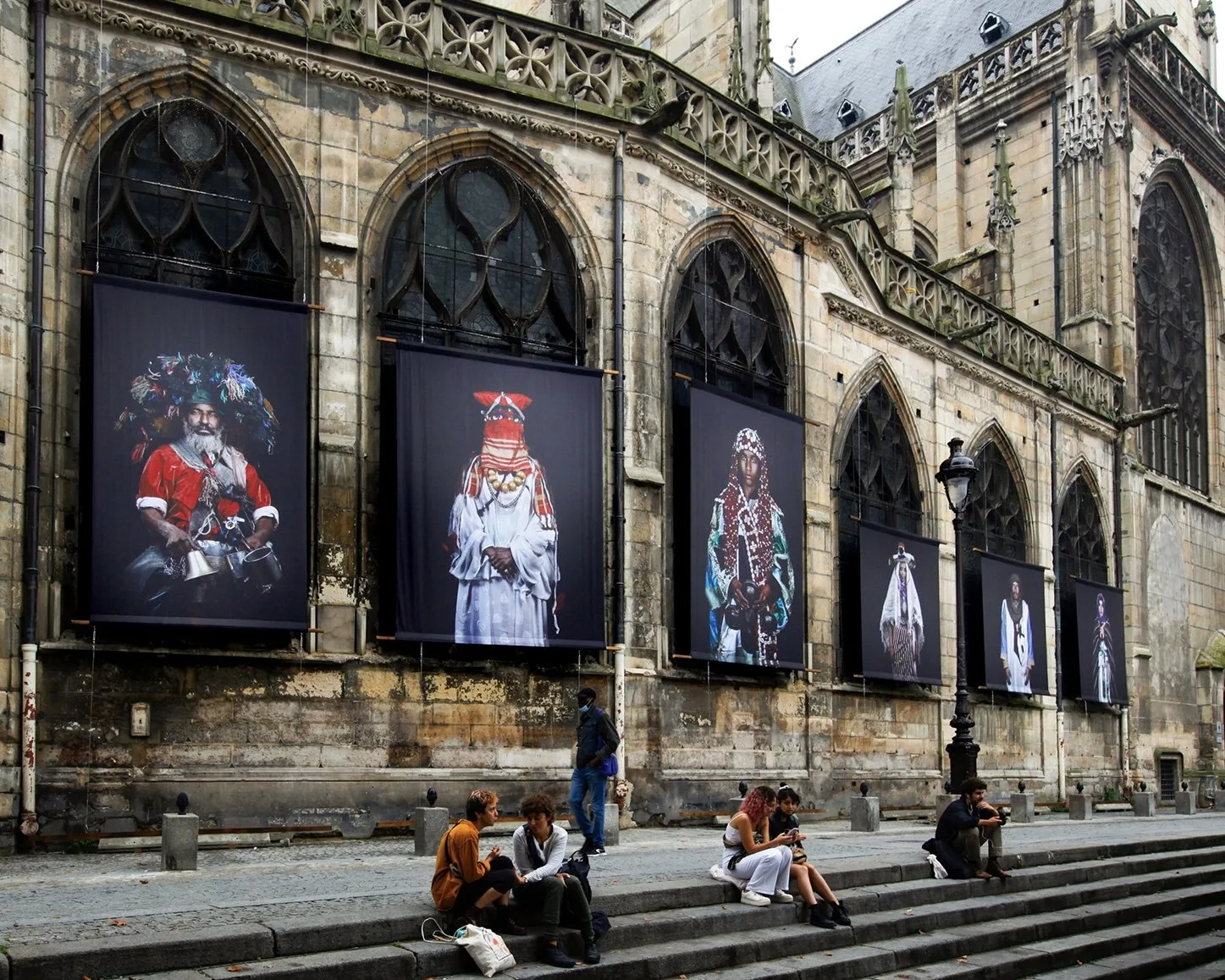Grands portraits de Leila Alaoui sur fond noir accrochés dans les arcs d'une église gothique ; des passants assis sur les marches.