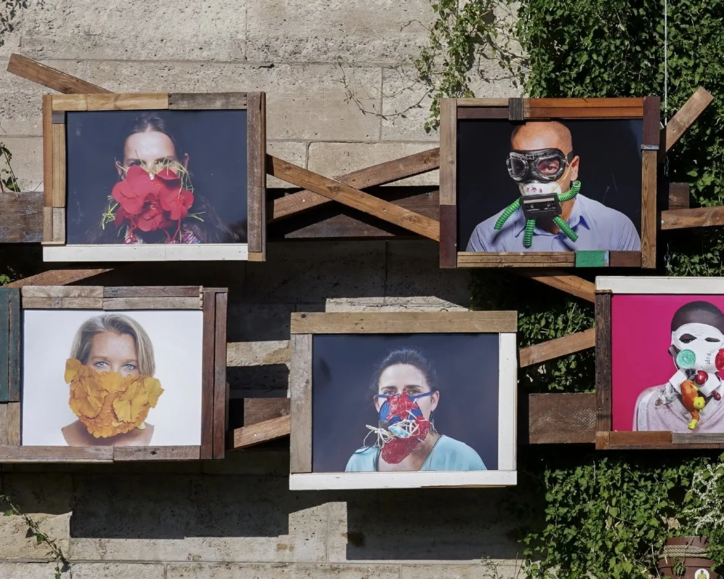 Portraits de personnes aux masques créatifs, dans des cadres en bois récupéré, accrochés à un mur de pierre sur les quais de Seine.