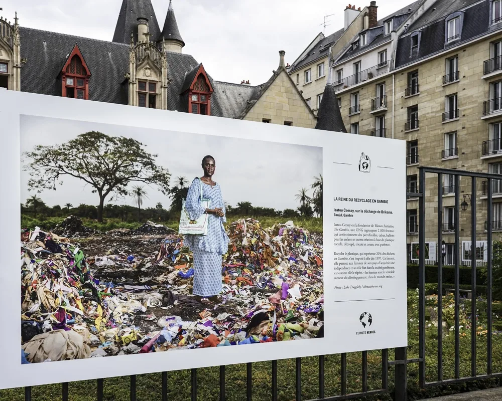 Panneau Climate Heroes contre une grille : une femme debout sur une décharge de déchets colorés, devant des immeubles du quartier Saint-Paul.