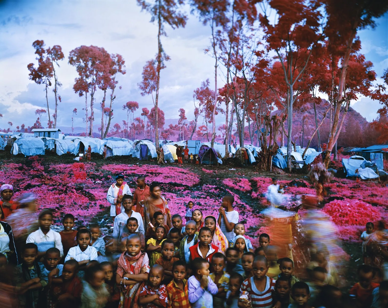 Foule d'enfants devant un camp de tentes bleues, la végétation virée au rose et au rouge par le film infrarouge de Richard Mosse.