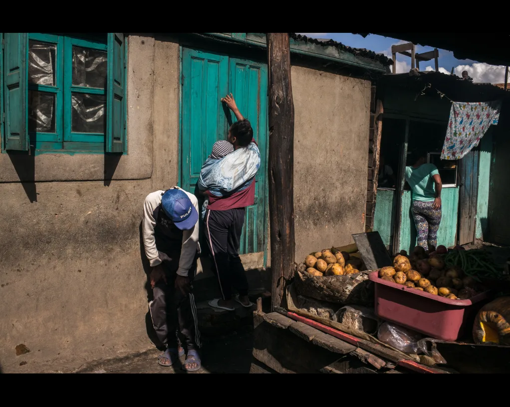 Rue à Madagascar : une femme portant un bébé au dos lève le bras vers une porte verte, un homme penché, des pommes de terre sur un étal.