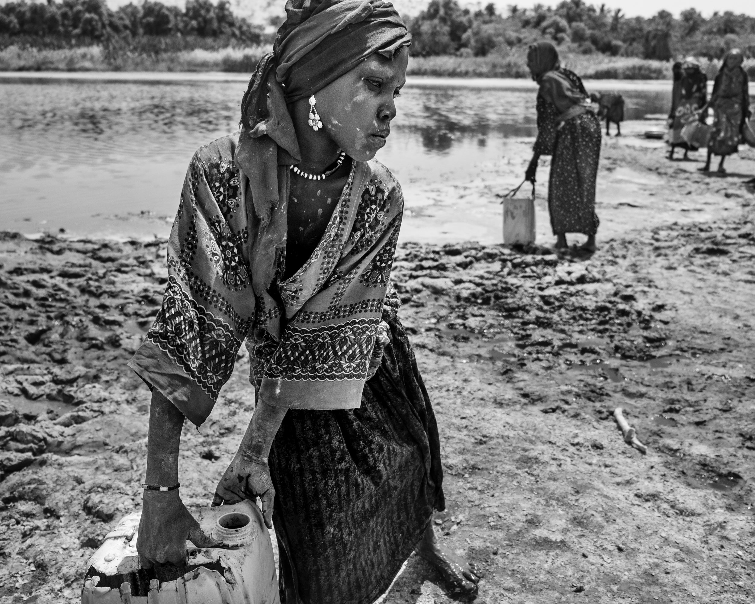 En noir et blanc, une femme tire un bidon en plastique sur une rive boueuse, d'autres femmes puisent de l'eau derrière elle.