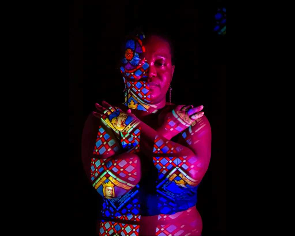 Portrait d'une femme aux bras croisés sur la poitrine, le corps couvert de la projection colorée d'un vitrail à figures couronnées.