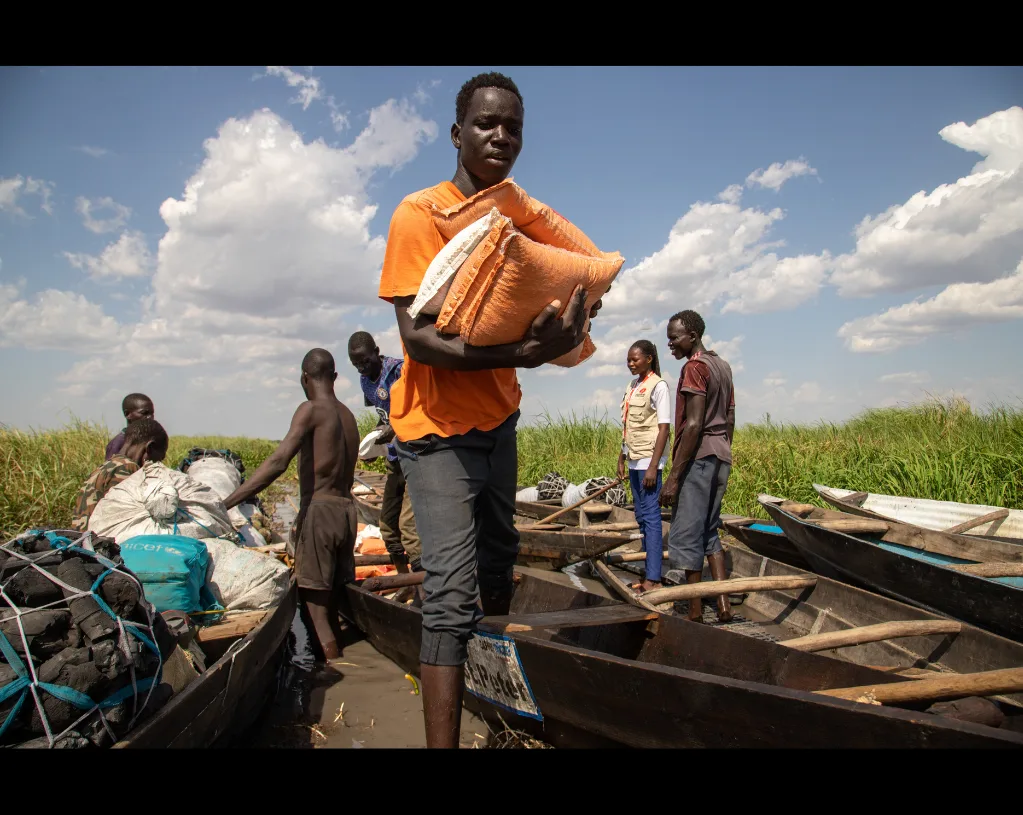 Au Soudan du Sud, un jeune homme en tee-shirt orange porte un grand sac sur l'épaule, entre des pirogues amarrées dans les herbes.
