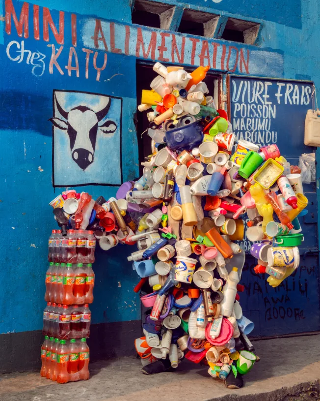 Personnage couvert de gobelets, bouteilles et bidons en plastique devant une épicerie peinte en bleu, « Homo Detritus » de Stephan Gladieu.