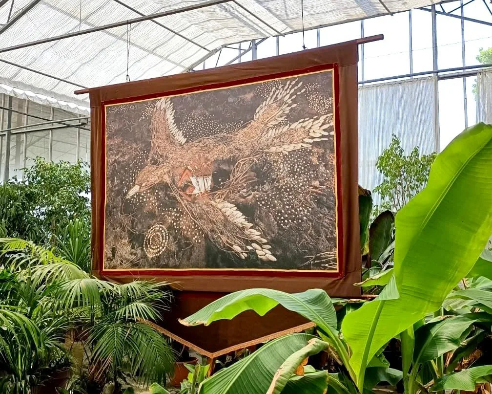 Un grand tirage bordé de tissu, montrant un oiseau aux ailes déployées, suspendu dans une serre parmi des bananiers.