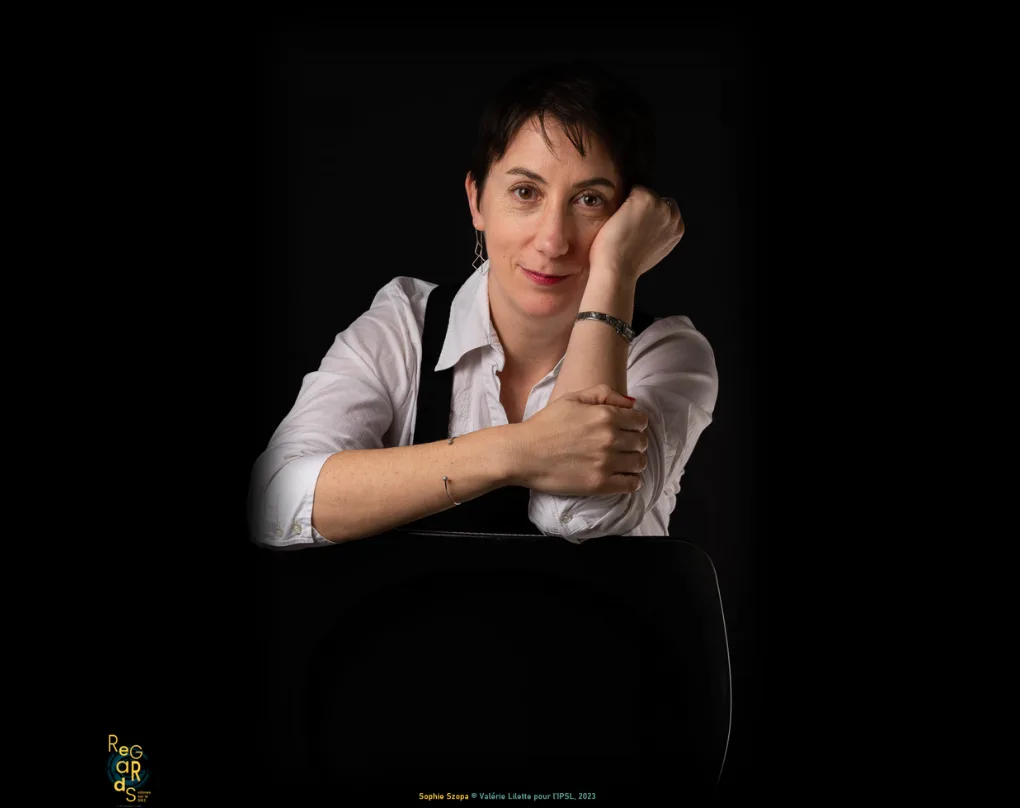 Portrait d'une femme aux cheveux courts, en chemise blanche, accoudée au dossier d'une chaise, la tête sur le poing, sur fond noir.