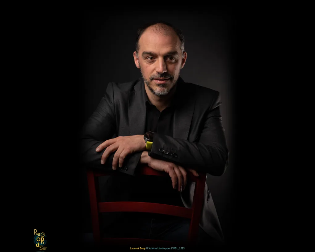 Portrait d'un homme à la barbe courte, en veste noire, accoudé au dossier d'une chaise rouge, sur fond noir.