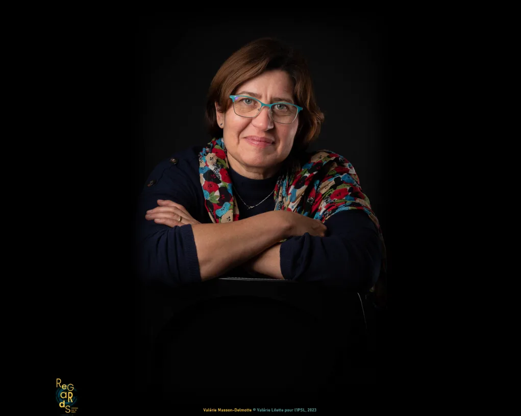 Portrait d'une femme souriante aux lunettes turquoise et au foulard fleuri, bras croisés sur le dossier d'une chaise, sur fond noir.