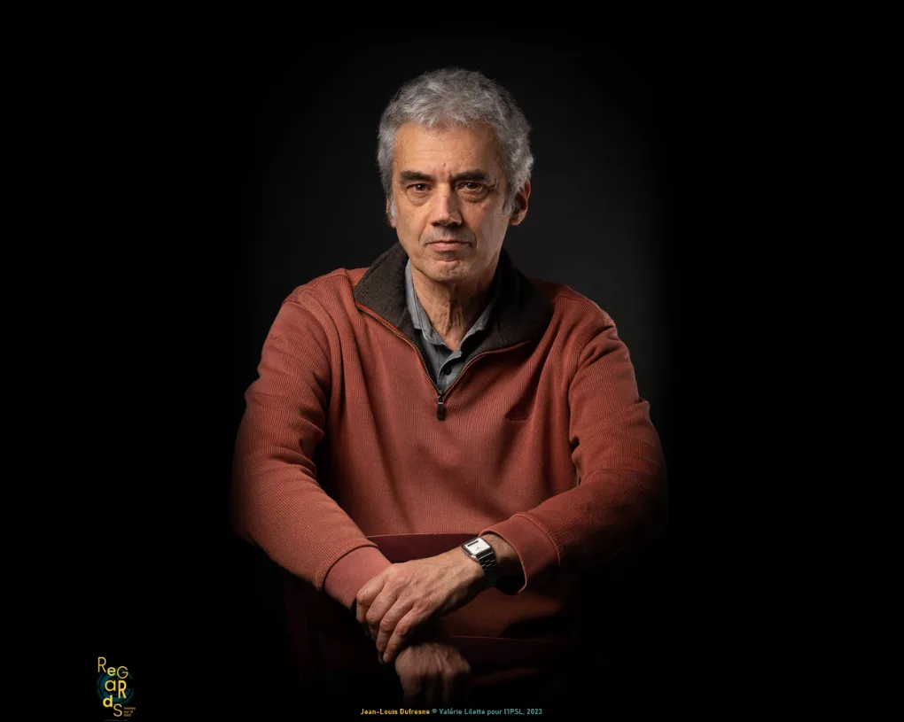 Portrait d'un homme aux cheveux gris, en pull zippé rouge brique, assis les mains croisées sur le dossier d'une chaise, sur fond noir.