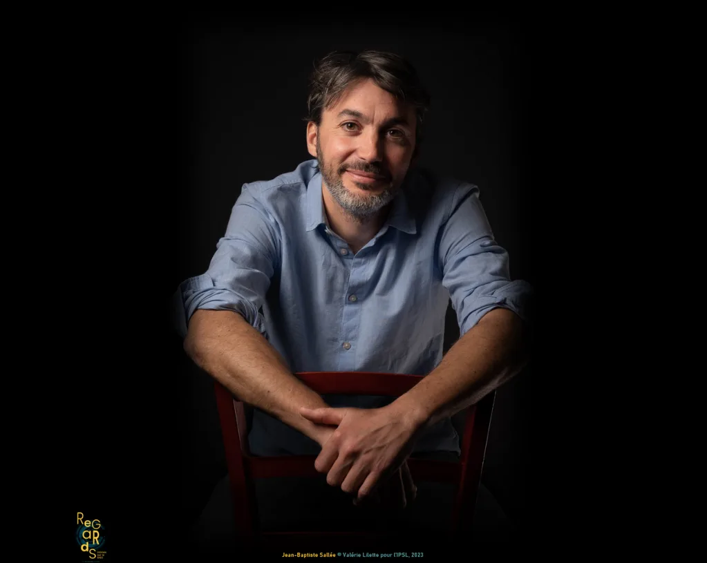 Portrait de Jean-Baptiste Sallée en chemise bleu ciel, assis à califourchon sur une chaise rouge, souriant, sur fond noir.